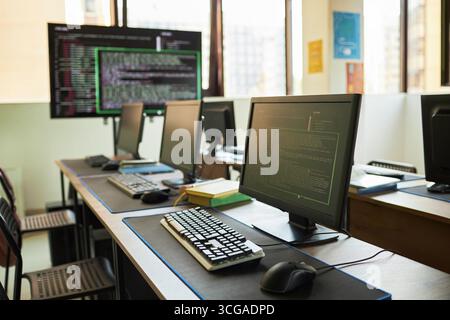 Empty computer lab featuring multiple desktop monitors displaying programming code on screens, keyboards and mice arranged on desks, large window providing natural light, no people present Stock Photo