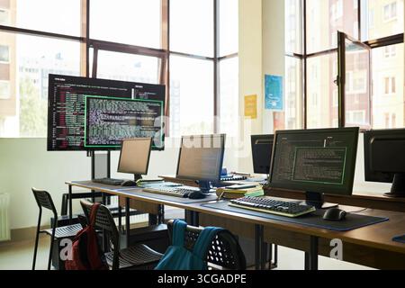 Empty classroom with multiple computer workstations displaying programming code on monitors, modern educational setting with large windows and backpacks on chairs Stock Photo
