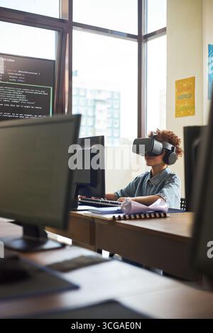 Black teenager wearing virtual reality headset sitting at desk using computer in modern classroom, engaging with coding software on monitor, surrounded by multiple computer screens Stock Photo