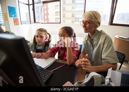 Caucasian middle aged woman assisting two girls, one Caucasian and one possibly Hispanic, working together at computer in modern classroom, both girls appearing to be children Stock Photo