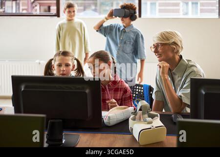 Caucasian senior woman supervising two preteen girls working at computer while two boys in background using virtual reality headset in modern classroom technology lesson Stock Photo