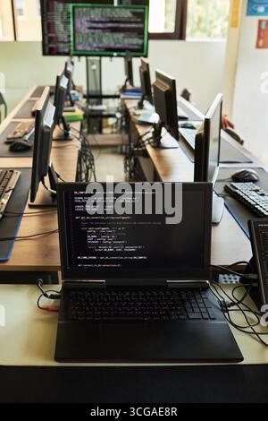 Row of computers on desks in modern office with open laptop displaying programming code in foreground, multiple monitors and empty chairs visible in background, no people present Stock Photo