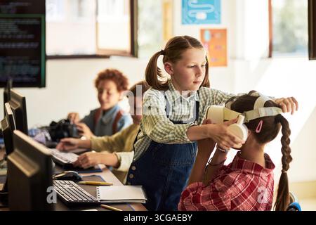 Caucasian girl helping another Caucasian girl put on virtual reality headset in classroom while two multiethnic boys using computers in background, children engaging with technology together Stock Photo