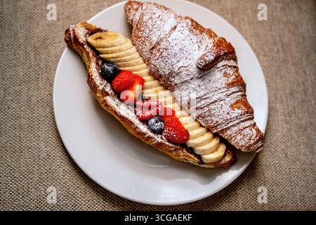 Sweet croissant with fruit (strawberry,banana and raspberry) with chocolate on white plate on beige fabric. Stock Photo