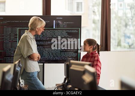 Middle aged Caucasian woman standing and talking with preteen girl in computer classroom, large monitor displaying programming code in background, both engaging in discussion Stock Photo