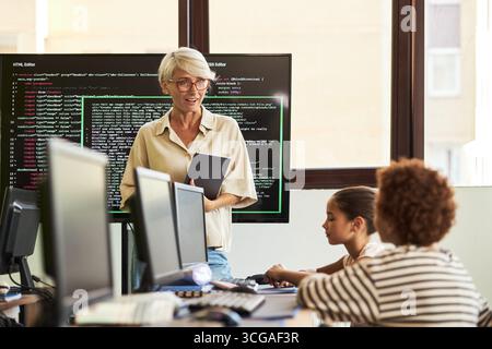 Middle aged Caucasian woman teaching coding class, holding digital tablet while standing near large monitor with code, two diverse children sitting at desks using computers, classroom setting Stock Photo