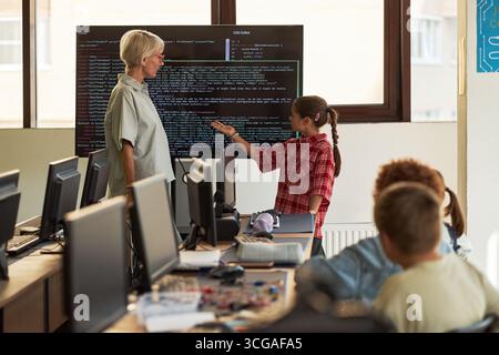 Senior Caucasian woman standing near large monitor displaying code, interacting with preteen Caucasian girl presenting programming project in computer classroom with students watching Stock Photo