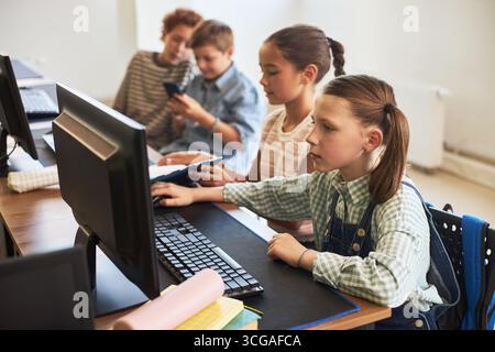 Group of Caucasian and multiethnic children sitting at desk using desktop computers and smartphone in classroom, focused expressions showing teamwork and learning technology skills together Stock Photo