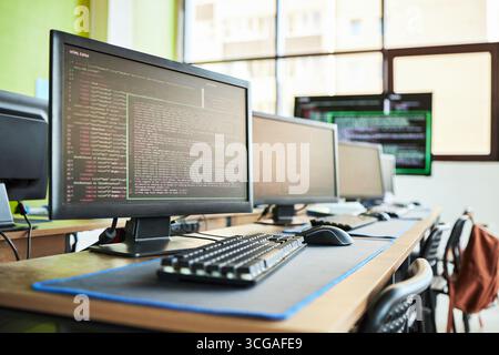 Row of desktop computers displaying programming code on monitors in modern computer lab, empty chairs lined up along desks, keyboards and mice arranged neatly, bright window background Stock Photo
