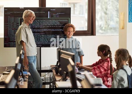 Senior Caucasian woman standing near digital screen displaying code, interacting with multiethnic group of children sitting at desks using desktop computers in classroom setting Stock Photo