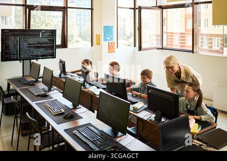 Caucasian female teacher assisting group of children using desktop computers in classroom, children sitting at desks working on coding tasks, large monitor displaying programming code Stock Photo