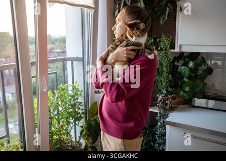 Thoughtful man holding cat while gazing out window on quiet day off in peaceful solitude, slow life Stock Photo