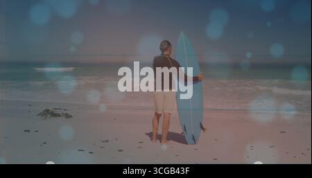 Mature man wearing black rash guard standing on sandy beach with blue surfboard and pebbles Stock Photo