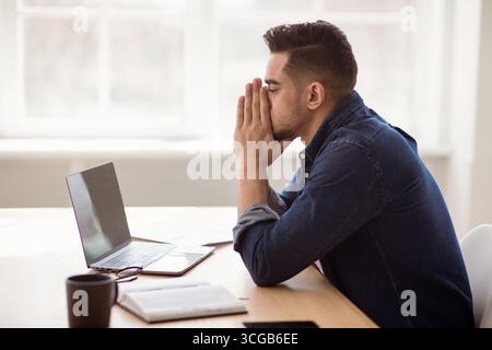 Exhausted arab man sitting in front of laptop, office interior Stock Photo