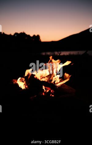 Campfire flames burning brightly with mountains and lake silhouette at sunset. Stock Photo