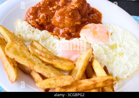 Meatballs simmering in rich tomato sauce, accompanied by two perfectly fried eggs and crispy chips, presented on a white plate Stock Photo