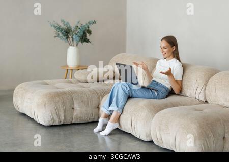 Puzzled lady in casual looking at laptop screen and raising hands up, sitting on couch at home Stock Photo