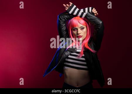Fashion forward woman with vibrant pink hair and striped crop top poses in dramatic scene. Stylish female model with arms above head, confidently stands in artistic studio lighting. Stock Photo