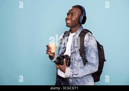 Male photographer poses with coffee cup and camera in hand, listening to music and carrying a backpack. Cheerful man wearing headphones stands against isolated backdrop, ready for photography session. Stock Photo