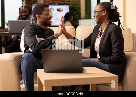 Two cheerful colleagues sitting on cozy sofa, high fiving, marking completion of collaborative business task. Young male and female employees in modern office, celebrating successful team effort. Stock Photo