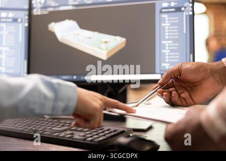 Selective focus on multiethnic developers with a clipboard and pen at desk, discussing prototypes. They are collaborating on design adjustments with CAD software displayed on a desktop pc monitor. Stock Photo