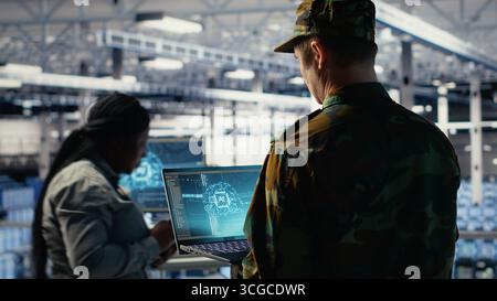 Soldier in engineering role uses AI application on laptop to manage server hub systems. Army commander reviews secret intelligence information using artificial intelligence in data center, camera B Stock Photo