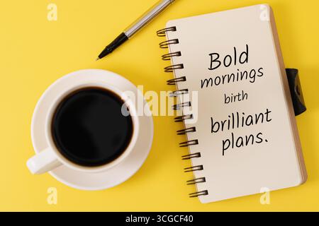White coffee cup, silver pen, and open notebook with motivational text on a yellow background with copy space. Stock Photo
