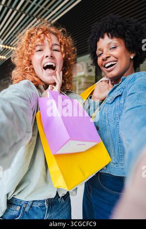 Two joyful women capturing a delightful selfie together while showcasing their colorful shopping bags filled with exciting purchases and enjoying Stock Photo