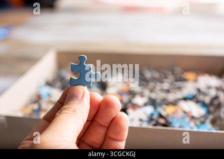 A closeup view of a hand holding a blue colored puzzle piece in front of a jigsaw puzzle box. Stock Photo