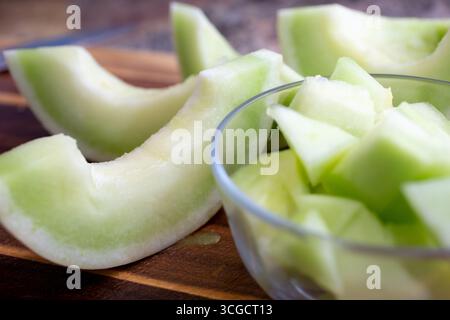 A view of a glass bowl filled with chopped honeydew melon, also featuring slices of honeydew adjacent, on a wooden cutting board. Stock Photo