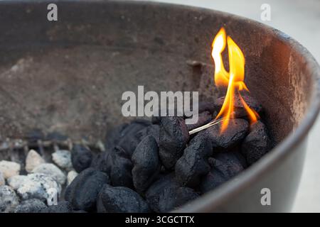 A closeup view of a pile of black charcoal on the side of a kettle barbecue grill, featuring a lit match igniting a flame. Stock Photo