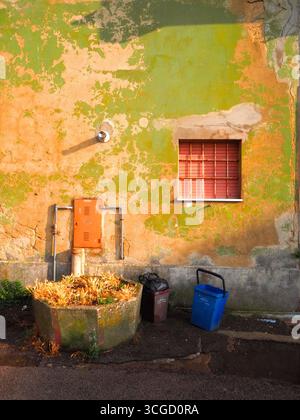 Enna wall with peeling green and orange paint illuminated by warm sunset light, red shuttered window, trash bins and small planter in rustic Sicilian Stock Photo