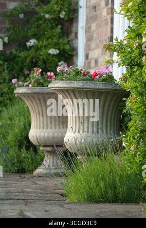 Two elegant stone planters filled with colorful flowers stand beside a brick wall, surrounded by lush greenery in a peaceful garden setting. Stock Photo