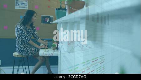Working analyst wearing dress sitting on stool at desk, using laptop with coffee mug and charts Stock Photo