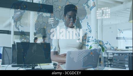 Working data analyst typing on silver laptop in open-plan office, with digital world map overlay Stock Photo
