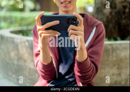An asian man is watching videos or playing games on smartphone holding horizontally with both hands while sitting in the park. Chilling Outdoor. Stock Photo