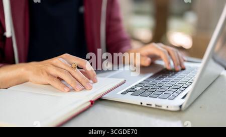 Close up of a hand holding pen resting on an open notebook and another hand typing on laptop while sitting at the table. Study or Working Outdoor. Stock Photo