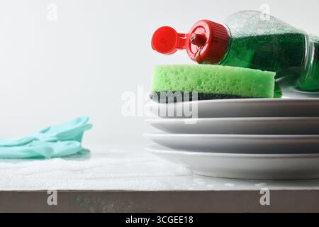 Detail of stack of white plates and green scouring pad and bottle with dishwashing product on white bench with water and soap and green gloves on isol Stock Photo