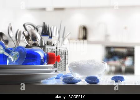 Detail of dishwasher products on white table like detergent in pods, salt in a bowl and rinse aid in a bottle on dishes with cutlery and glass, and wh Stock Photo