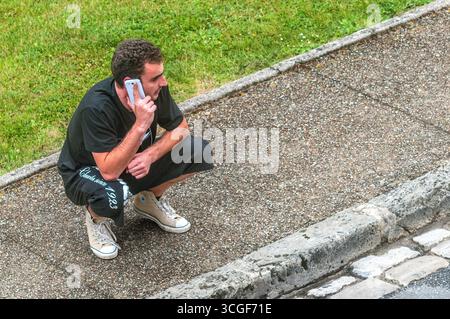 Overhead view of young man holding smartphone whilst crouching on pavement - Preuilly-sur-Claise, Indre-et-Loire (37), France. Stock Photo