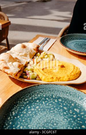 creamy curry hummus served with warm halved naan bread and a colorful fresh salad on a wooden table at a sunny sidewalk cafe surrounded by city life Stock Photo