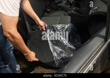 a man removes the protective plastic cover from a new black floor mat before placing it on the passenger side of a car interior, preparing to install Stock Photo