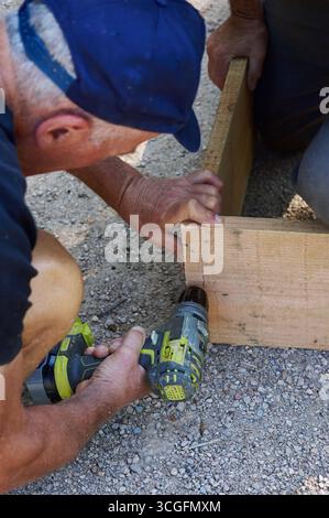 Close-up of senior man using power drill to assemble wooden crate, concept of manual work and craftsmanship. Stock Photo