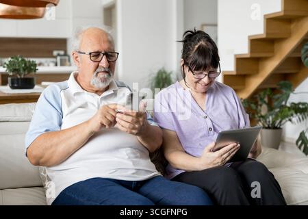 Elderly man and woman sitting on a couch, focused on smartphone and tablet, enjoying technology together in a cozy modern living room. Stock Photo