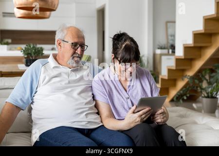 Smiling elderly couple sitting on a couch at home, using a tablet for entertainment and connection in a bright modern living room. Stock Photo