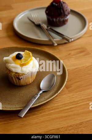 A delicious vanilla cupcake topped with white frosting, a blueberry, and a lemon slice on a rustic ceramic plate with a spoon, with a second cupcake Stock Photo