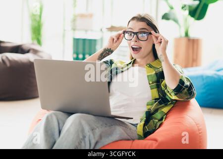 Freelancer woman wearing glasses using a laptop while seated in a cozy and modern interior, expressing delight and success. Stock Photo