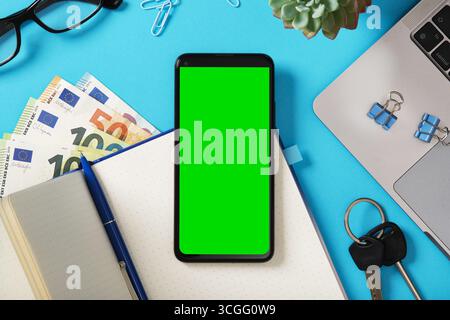 Smartphone mockup with green screen on desk next to laptop, notebook, euro banknotes, and stationery. Demonstration for your mobile app. Stock Photo