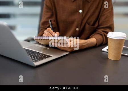 Man in a casual brown shirt jotting notes with a pen beside a laptop and coffee. Stock Photo