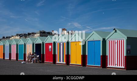 A couple sitting outside a colourful beach huts on Brighton seafront against a bright blue sky in August 2025. Stock Photo